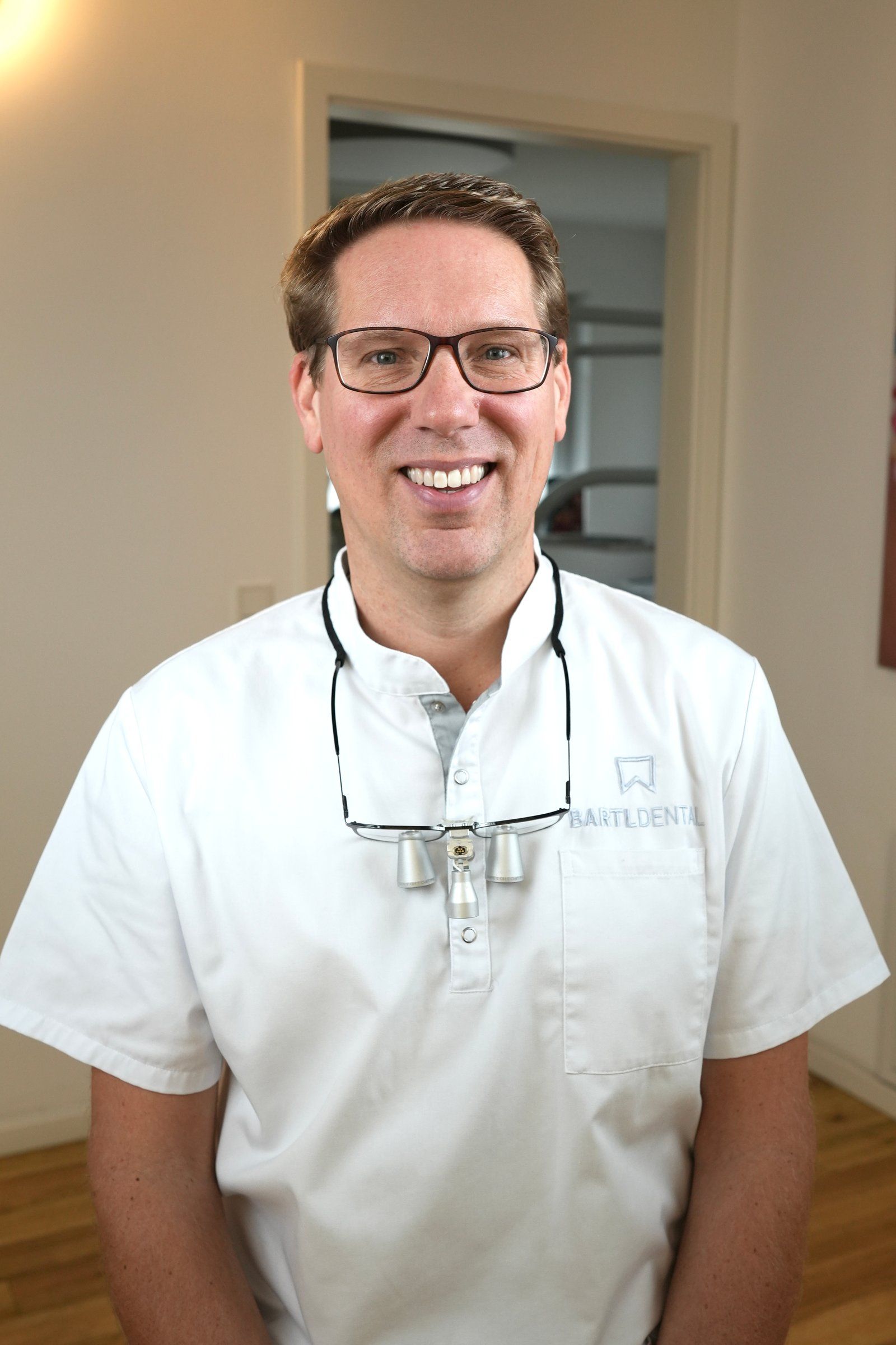 Zahnarzt M.Sc. Robert Bartl, Praxisinhaber mit Schwerpunkt Implantologie, lächelnd in weißer Praxiskleidung mit Lupenbrille vor einem Behandlungszimmer der Praxis in Bobingen