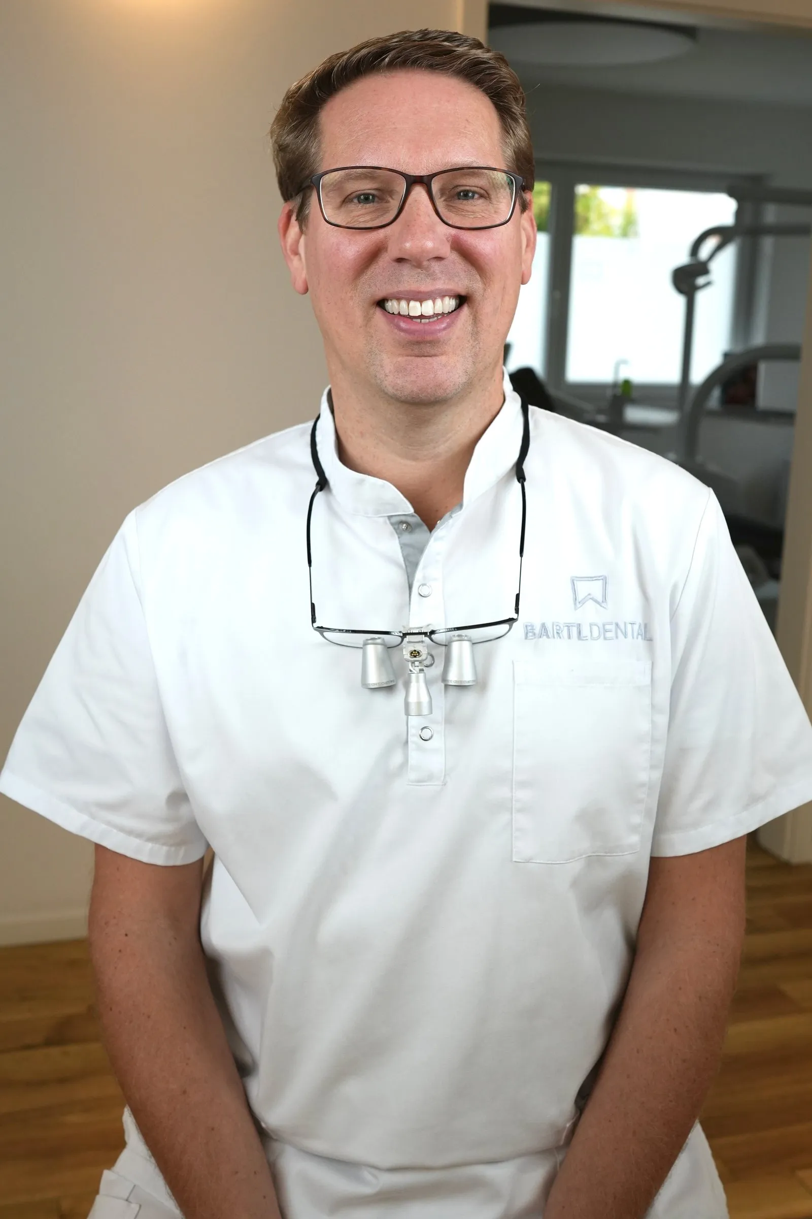 Zahnarzt M.Sc. Robert Bartl, Inhaber von Bartl Dental in Bobingen, sitzend und lächelnd in weißer Praxiskleidung mit Lupenbrille, hinter ihm ein Behandlungszimmer der Praxis