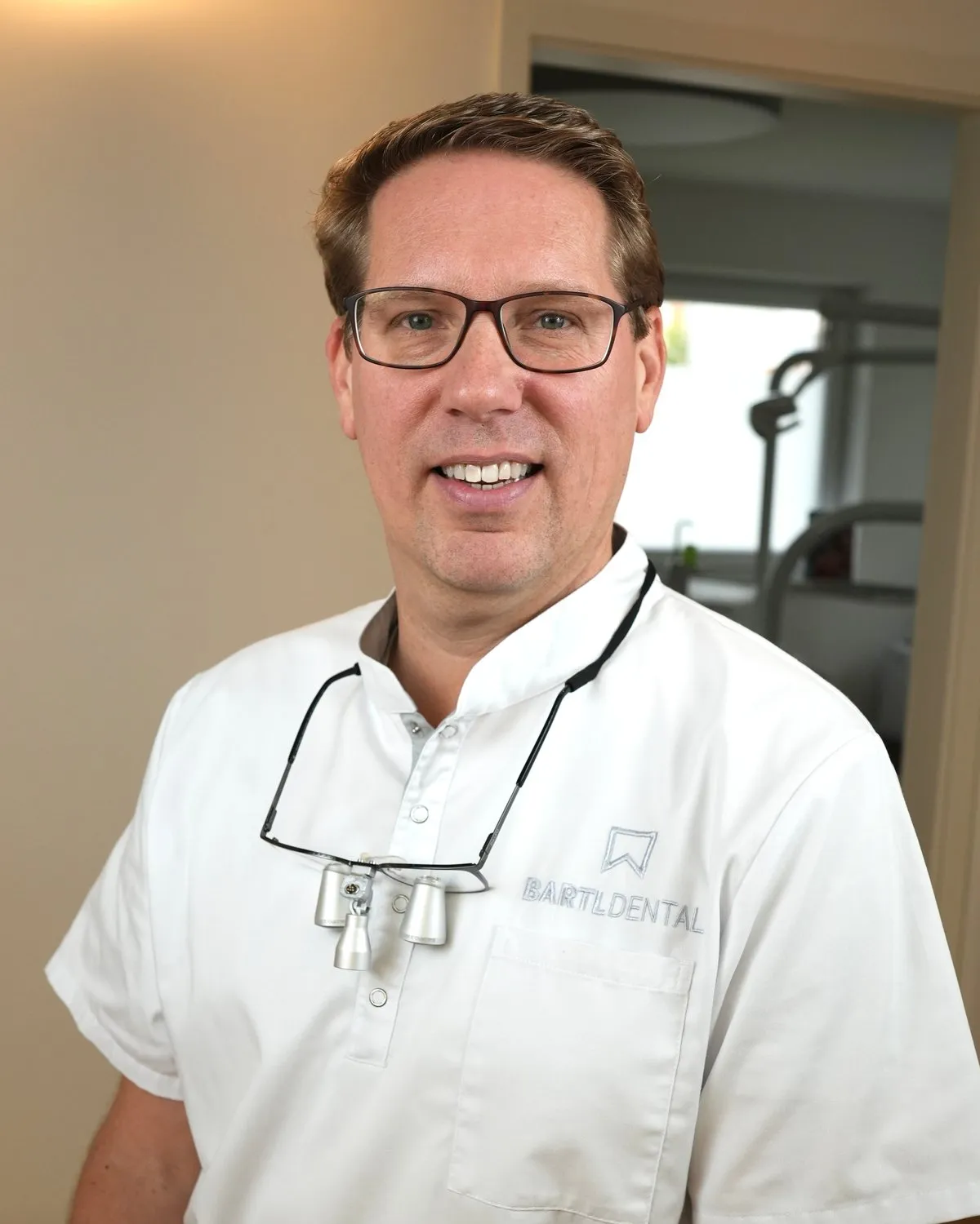 Zahnarzt M.Sc. Robert Bartl, Inhaber von Bartl Dental in Bobingen, lächelnd in weißer Praxiskleidung mit Lupenbrille
