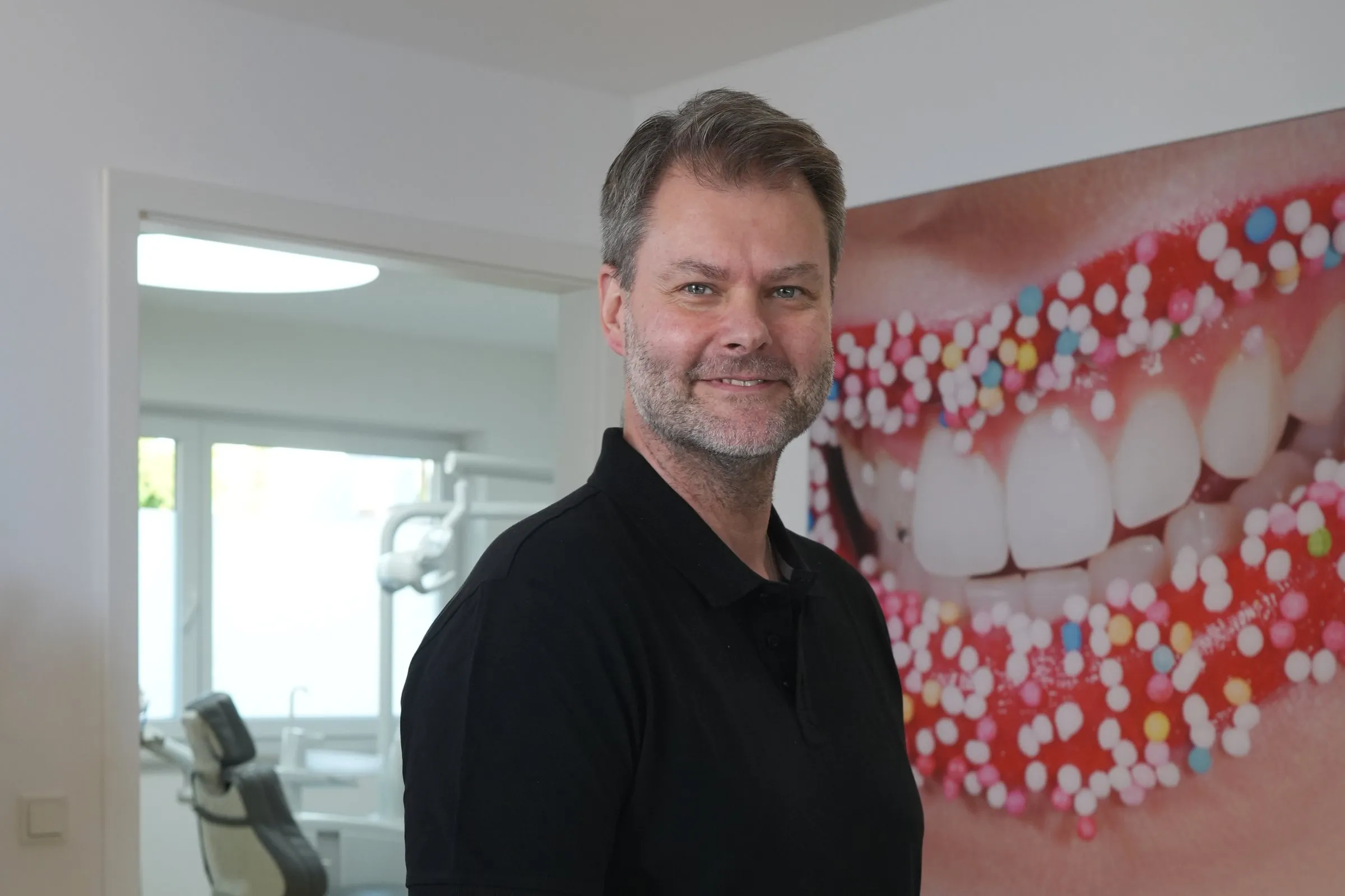 Zahntechniker David in schwarzem Poloshirt in der Praxis Bartl Dental, hinter ihm das Wandbild mit dem großen Lächeln