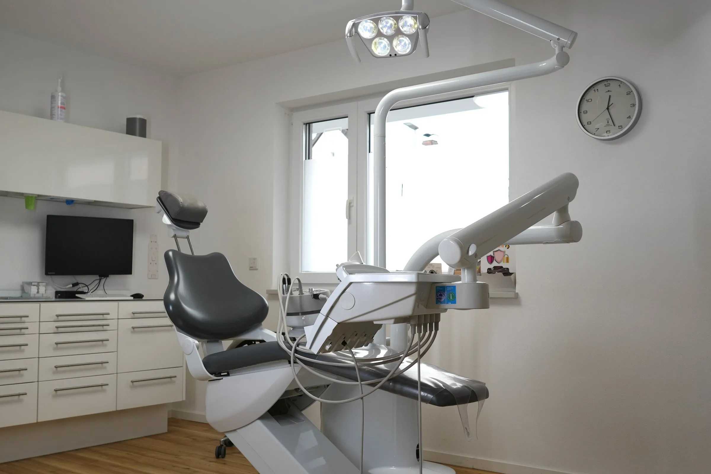 Behandlungszimmer mit Behandlungsstuhl und Leuchte vor dem Fenster, links weiße Schränke und ein Bildschirm, rechts eine Wanduhr