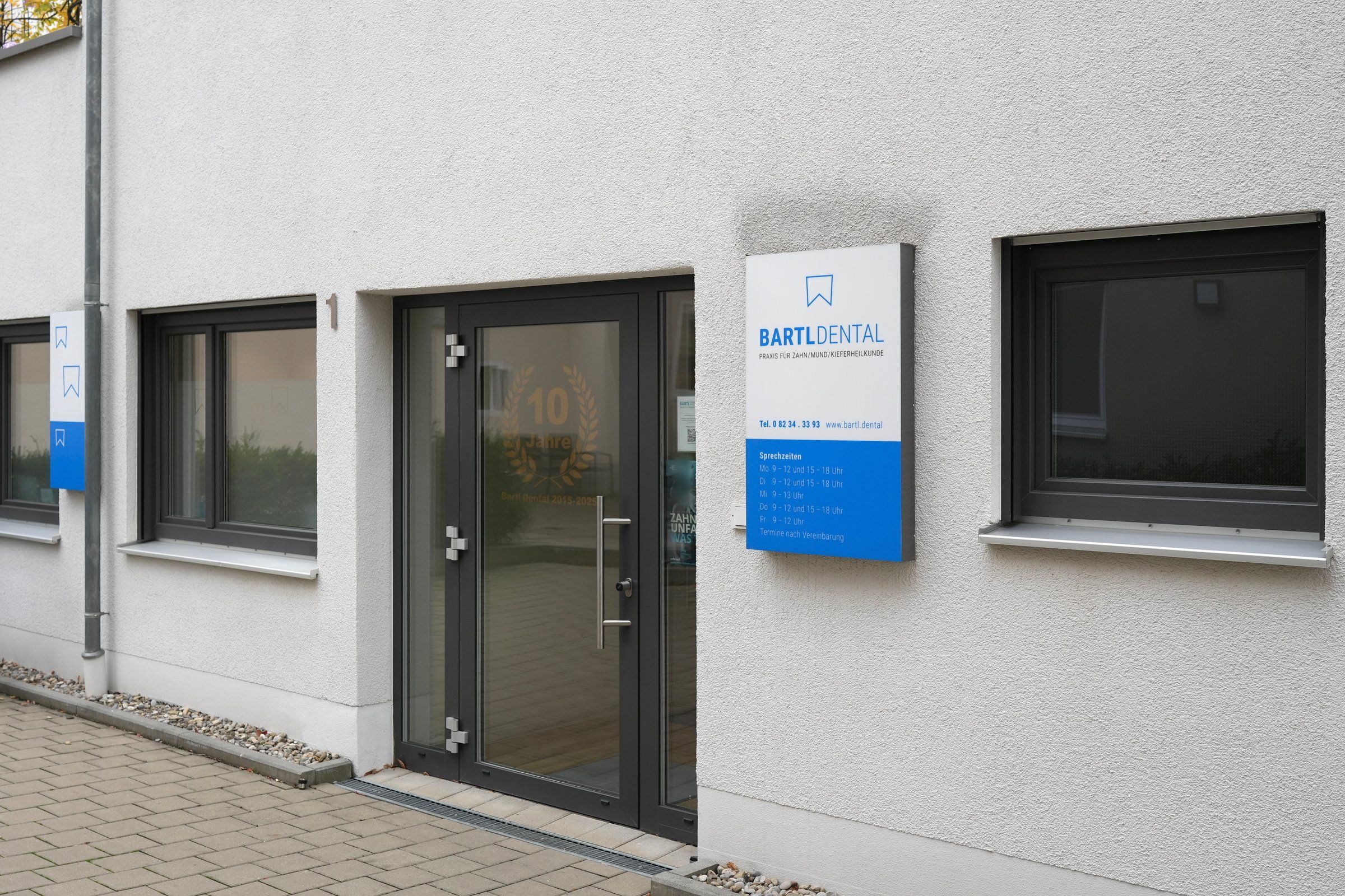 Eingang der Zahnarztpraxis Bartl Dental in Bobingen: ebenerdige Glastür mit dem Schriftzug „10 Jahre“, links daneben die Hausnummer 1, rechts das Praxisschild mit Telefonnummer und Sprechzeiten