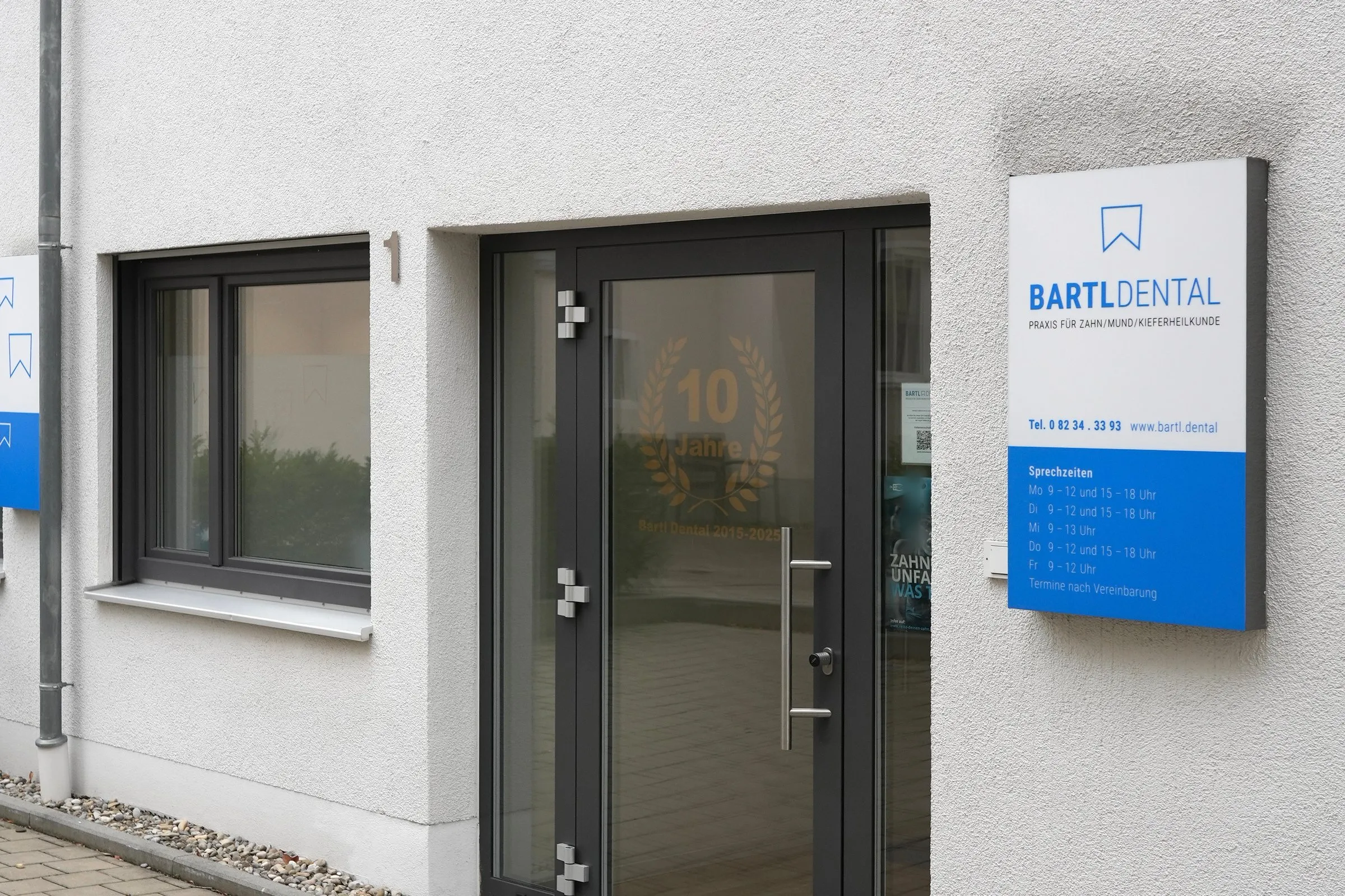 Glastür der Praxis Bartl Dental mit dem goldenen Schriftzug „10 Jahre“, rechts das weiß-blaue Praxisschild mit den Sprechzeiten