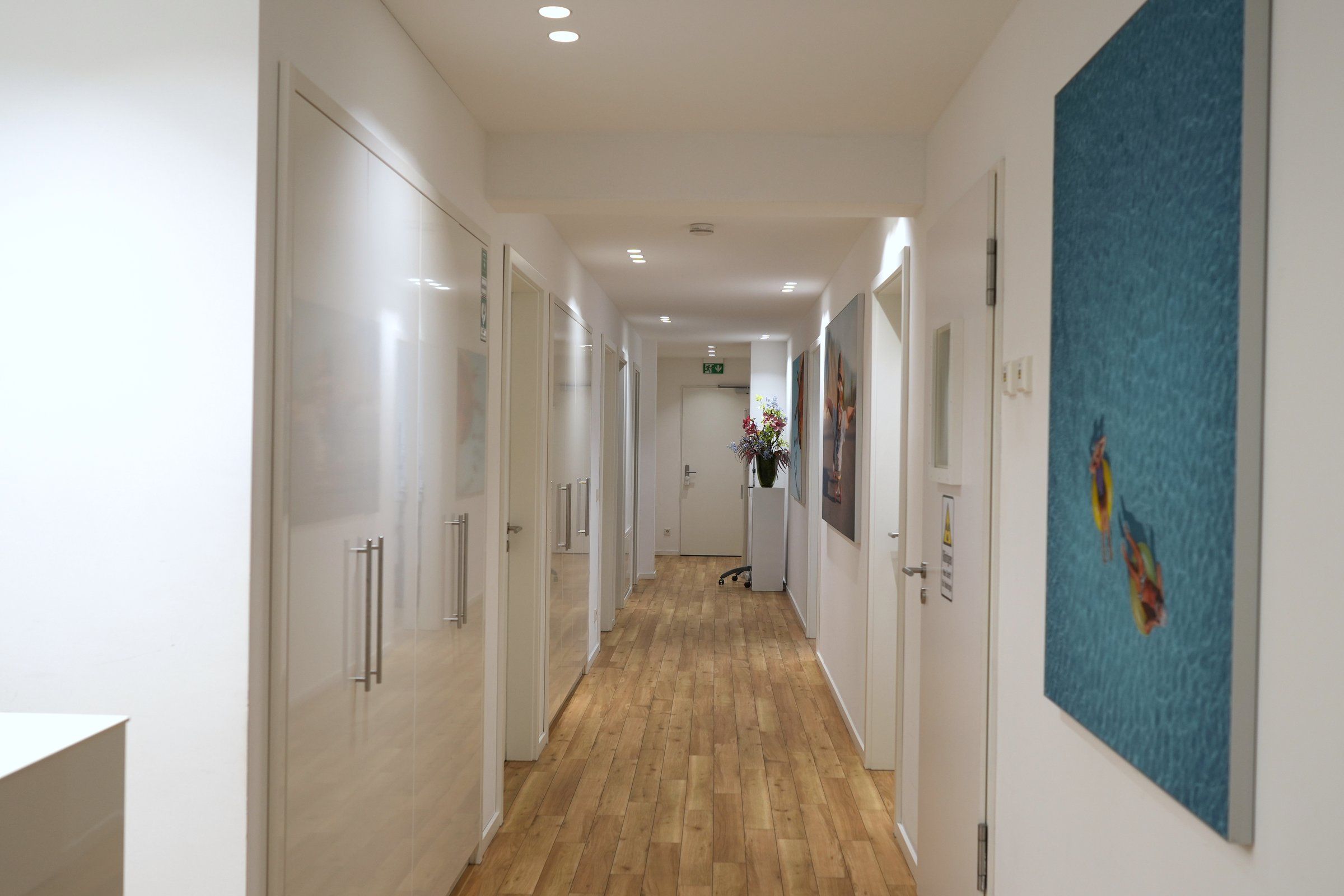 Langer Flur der Praxis Bartl Dental in Bobingen mit Holzboden und weißen Türen, rechts ein großes blaues Wandbild, am Ende ein Blumenstrauß