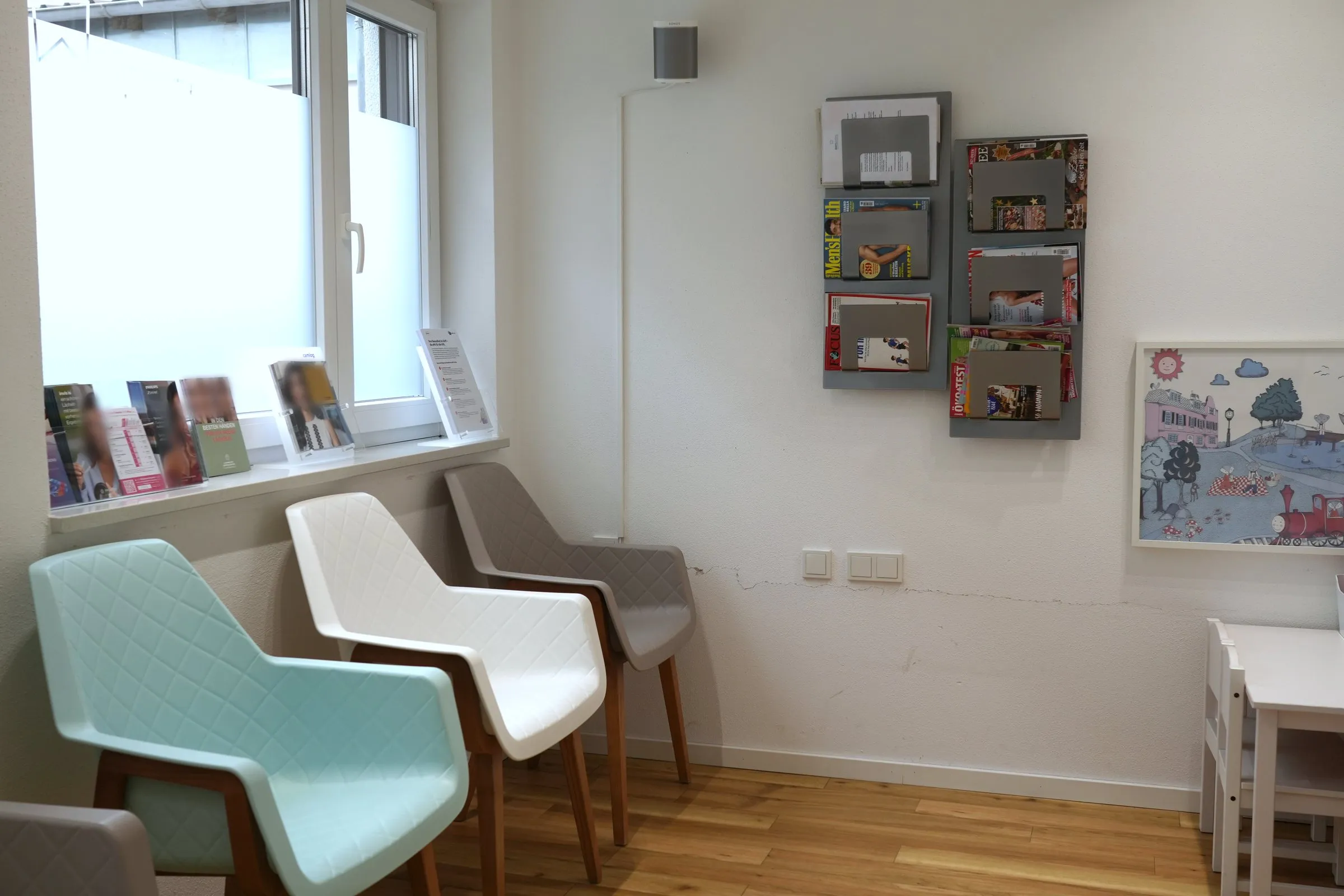 Wartezimmer von Bartl Dental: Stühle in Mint, Weiß und Grau unter dem Fenster, an der Wand ein Zeitschriftenregal und ein gerahmtes Kinderbild mit Eisenbahn, rechts ein Kindertisch